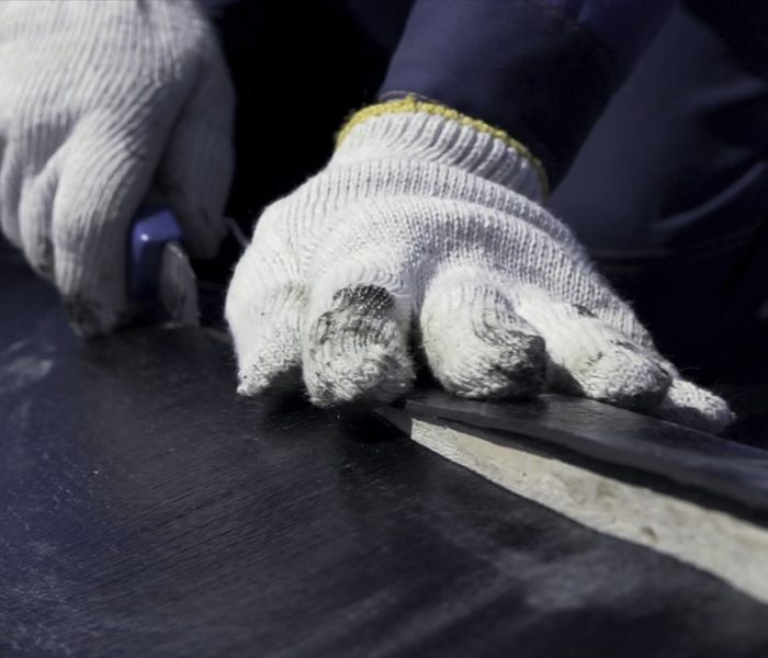 Close up for worker hands in white protective gloves cutting a black sheet of ruberoid with a